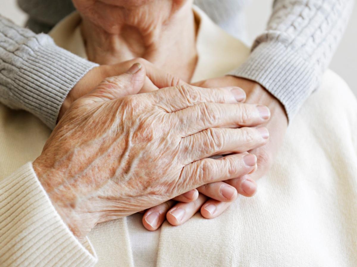 Die Hand einer Seniorin greift nach den helfenden Händen einer jüngeren Person.