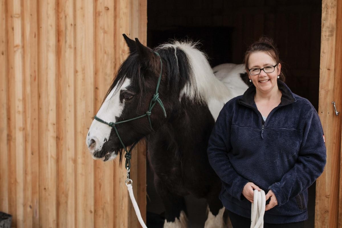 Tina Holzheimer lächelt in die Kamera, dabei hat sie die Zügel für ein Pferd, welches aus der Scheune kommt, in der Hand.