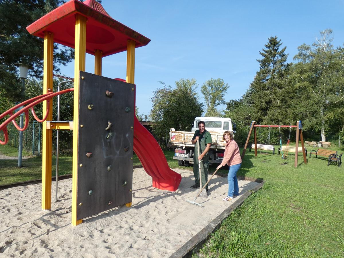 Mit einem Kleinlaster hat ein Mann Sand an eine Kinderrutsche gekippt. Er und eine Frau verteilen ihn mit Schaufel und Rechen überall am Spielgerät.