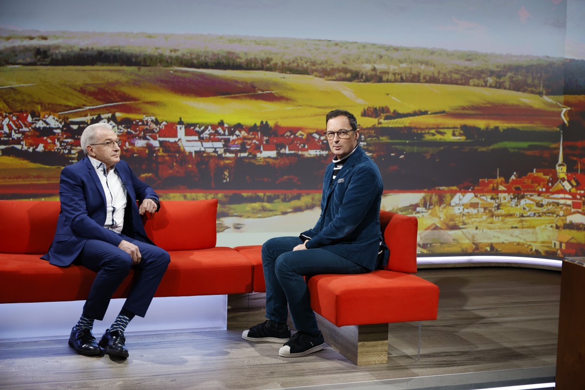 Zwei Männer sitzen in einem Fernsehstudio auf einem roten Sofa