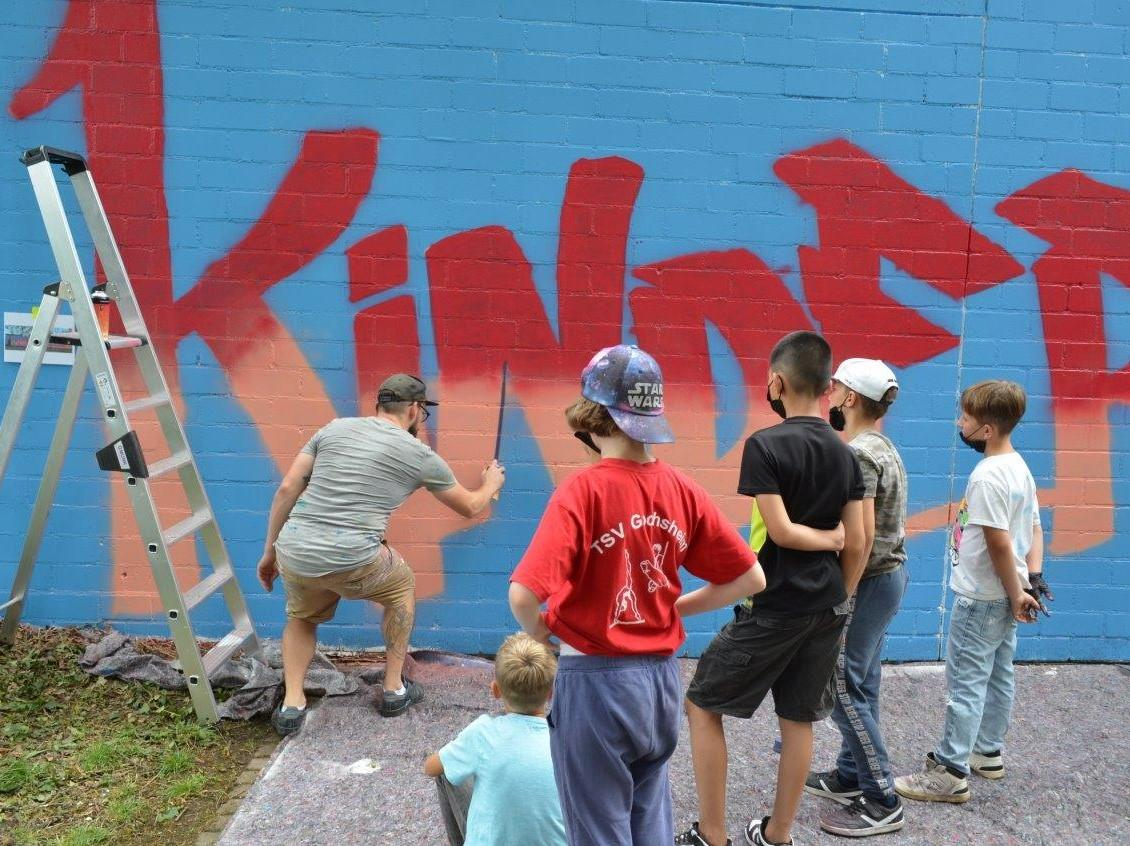 Graffitikünstler Christian Böhmer erledigt die Feinarbeit am Wort Kinder, während seine Helfer ihm über die Schulter schauen.