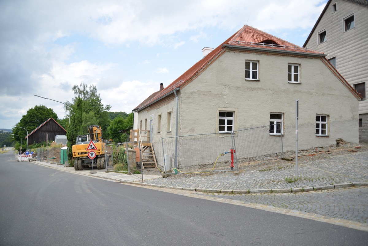 Das noch unverputze Bestandgebäude ist mit einem Bauzaun gesichert und befindet sich noch im Bau. Ein Bagger steht links daneben. Im Vordergrund ist die Straße des Ortseingangs zu sehen.