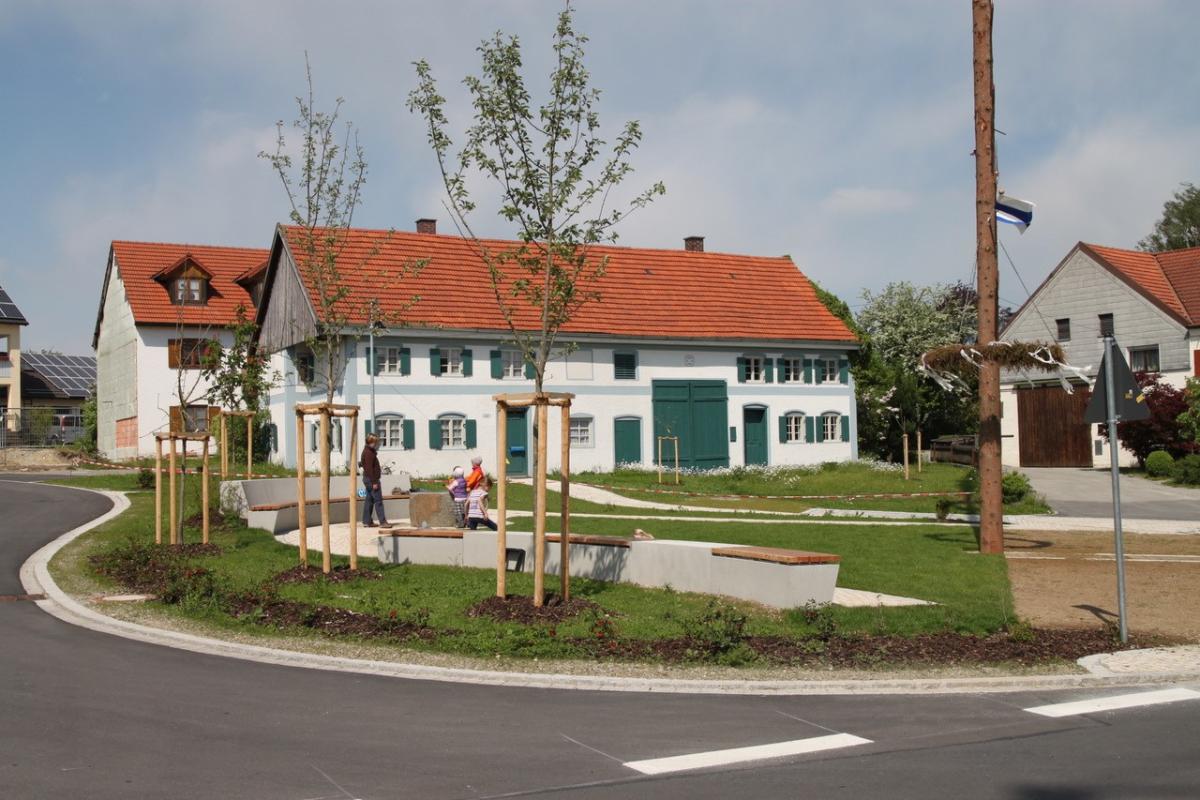 Bild von einem Dorfplatz mit einem Brunnen und einem Haus mit Fensterläden.