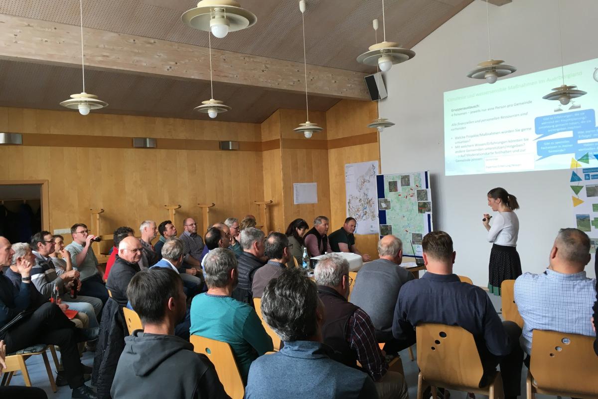 Ein Bild von einem Saal mit vielen Teilnehmern auf Stühle, vorne steht eine Frau, die etwas präsentiert.