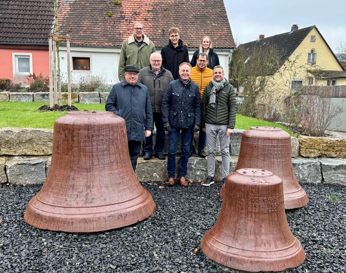 Eine Gruppe steht auf einem Platz hinter großen Kirchenglocken