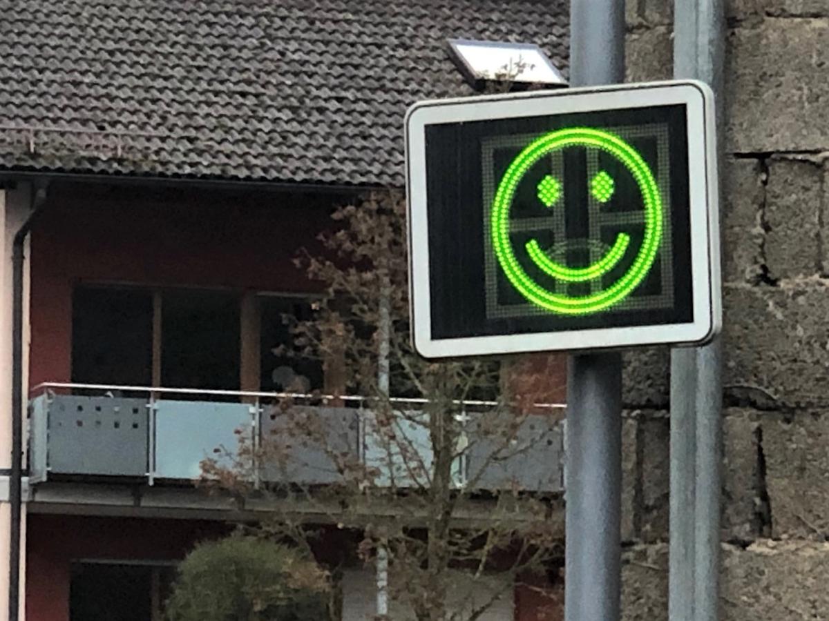 An einem Metallmasten hängt eine elektronische Geschwindigkeitsanzeigetafel. Sie zeigt zunächst das gefahrene Tempo und danach - wie hier - ein apfelgrünes Smiley (oder ein rotes Miesepetergesicht).